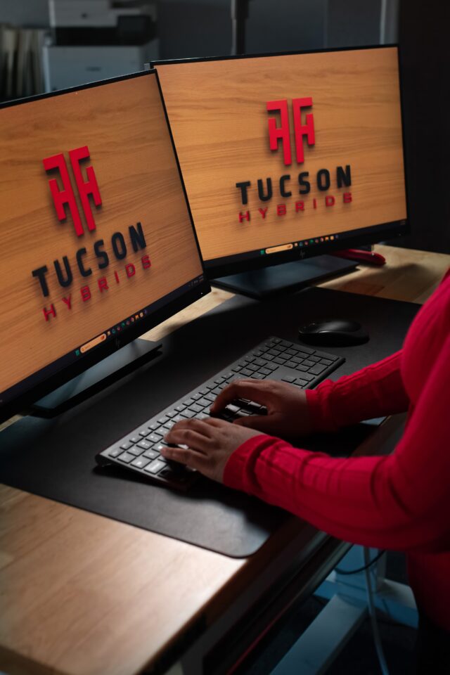 Service advisor at Tucson Hybrids working on dual monitors with company logo, handling hybrid and EV repair scheduling in Tucson, AZ auto shop specializing in battery diagnostics and electric vehicle services.